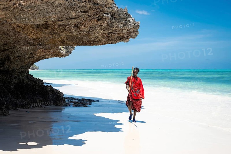 ZANZIBAR - OCTOBER 15, 2025: A Maasai man in traditional red clothing walks along a pristine white sand beach under a dramatic overhanging coral rock formation in Zanzibar, Tanzania