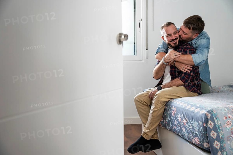 A joyful gay couple sitting on a bed, sharing a warm embrace. The intimate setting captures genuine happiness and affection, showcasing love and companionship in a cozy home environment