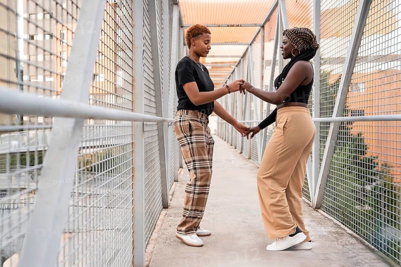 An affectionate LGBT multiethnic couple stands on a pedestrian bridge, engaged in a heartfelt moment. They are holding hands and smiling, representing love and diversity in an urban setting