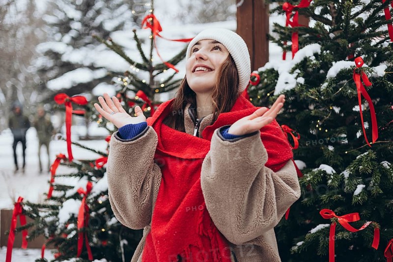 A young woman celebrates winter as she joyfully catches snowflakes near a festively decorated outdoor Christmas tree, dressed in cozy winter attire