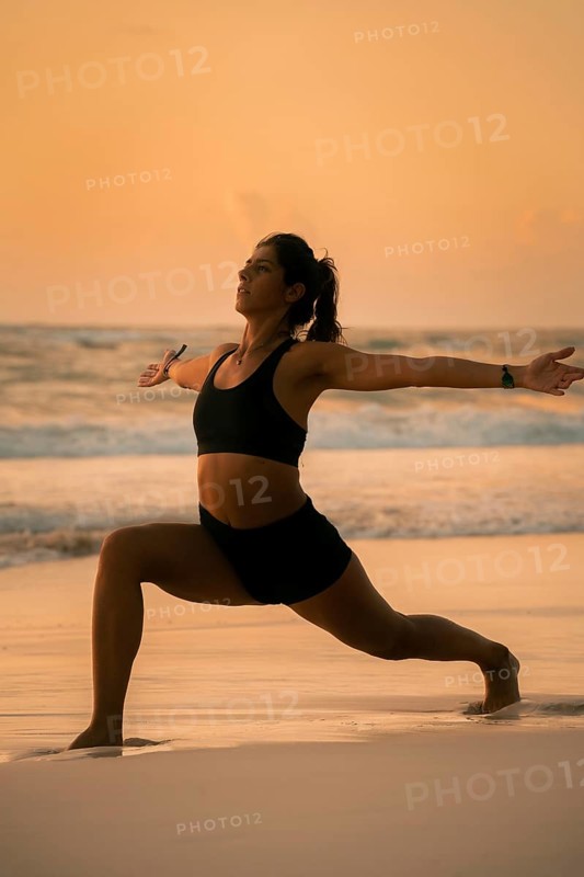 A serene image of a woman practicing yoga on a sandy beach at sunrise in Tulum, Mexico. She is in a strong warrior pose, embracing the peacefulness of the ocean and dawn light