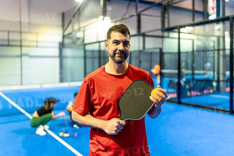 Adult male athlete wearing a red shirt holding a black carbon paddle and yellow ball while standing on a blue indoor pickleball court, preparing for a game
