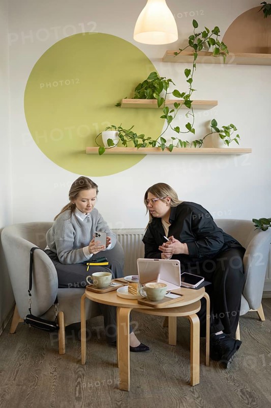 Two women engage in conversation in a cozy cafe setting. Surrounded by coffee, pastries, and greenery, they enjoy a relaxed and casual environment while using digital devices