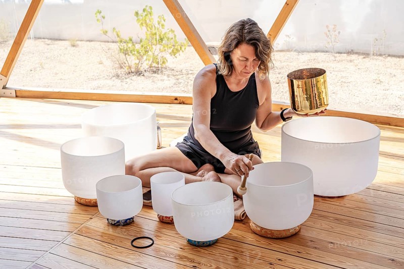 A woman is seated on a wooden floor, playing multiple crystal singing bowls. She gently taps them with a mallet, creating soothing sounds in a serene setting