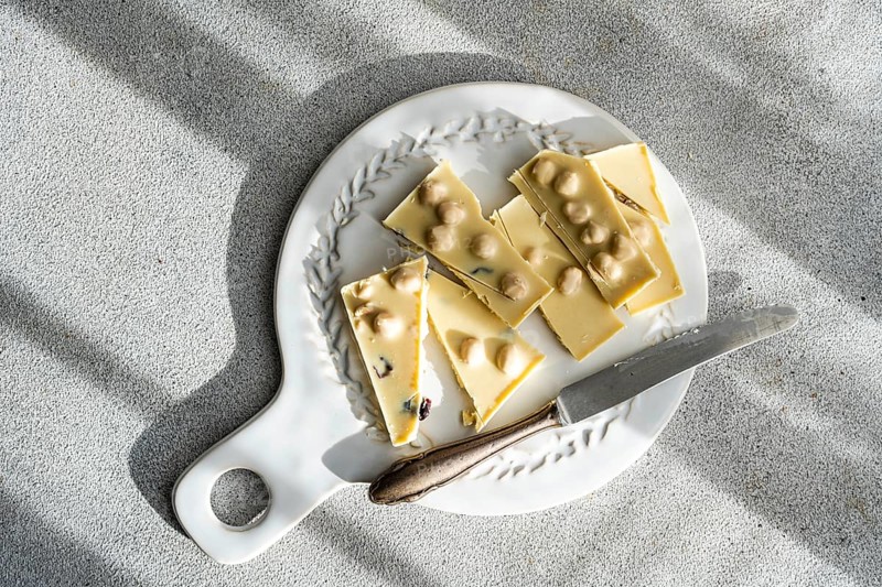 Top view of a sliced white chocolate bar with nuts, accompanied by a rustic knife on a decorative ceramic plate, beautifully illuminated by natural light against concrete background