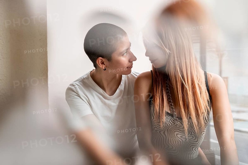 Intimate and candid capture of a lesbian couple sharing a tender moment in a softly lit room. They gaze into each other's eyes, reflecting love and connection