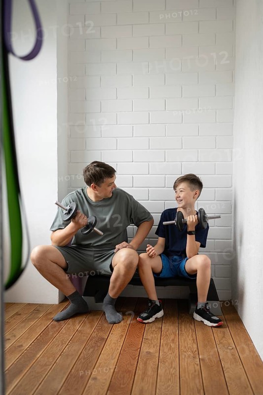 A father and son bond while exercising with dumbbells at home, sitting on a bench in a bright room. The interaction emphasizes fitness, unity, and healthy living