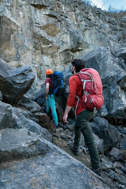Two hikers with backpacks approach a rocky cliff, preparing for a challenging climb. They are wearing helmets and climbing gear, ready for an outdoor adventure