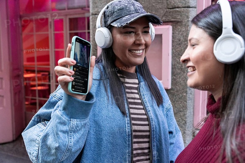 A joyful Latin lesbian couple, wearing headphones, shares a moment of connection while listening to music on a smartphone outdoors, surrounded by a vibrant urban setting