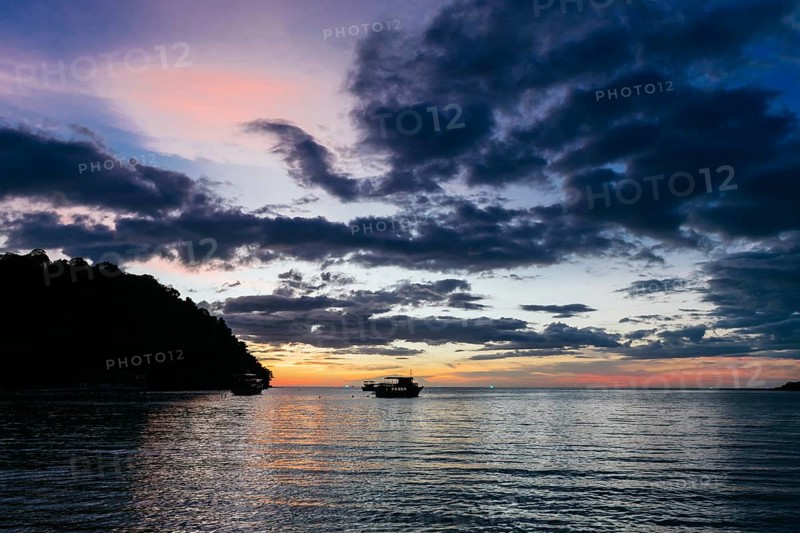 Traditional boats gently float on the tranquil waters of Koh Rong Island, Cambodia, as the sun sets, casting vibrant hues across the sky, creating a serene panorama