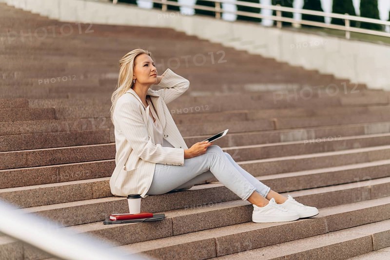 A businesswoman in casual attire sits thoughtfully on steps holding a tablet, with a coffee cup and notebook beside her. She appears relaxed in an outdoor urban setting