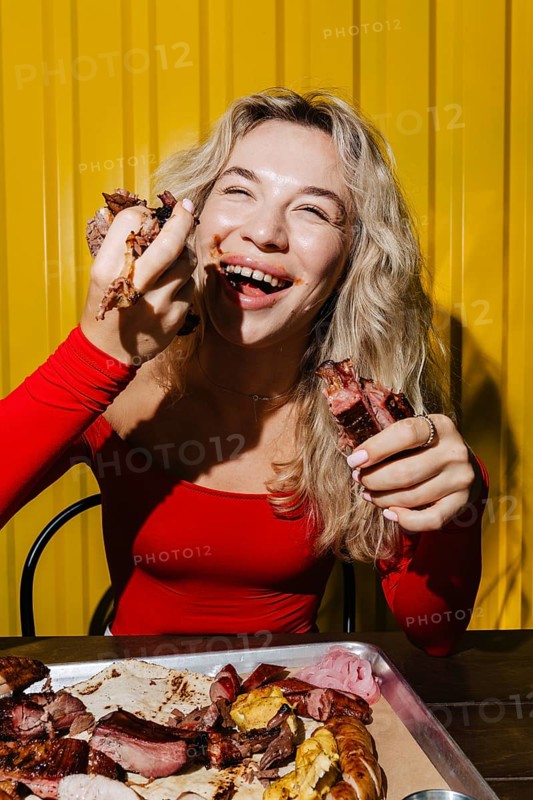 A happy woman in a red top enjoys eating barbecue meat with her hands. Sitting in front of a tray of grilled food, she smiles brightly, savoring the moment in a vibrant setting