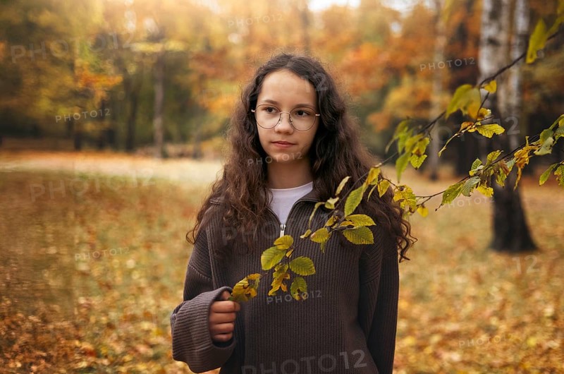 A young girl with glasses stands in an autumn setting, surrounded by vivid fall foliage. She is holding a branch with yellow leaves, capturing the essence of the season