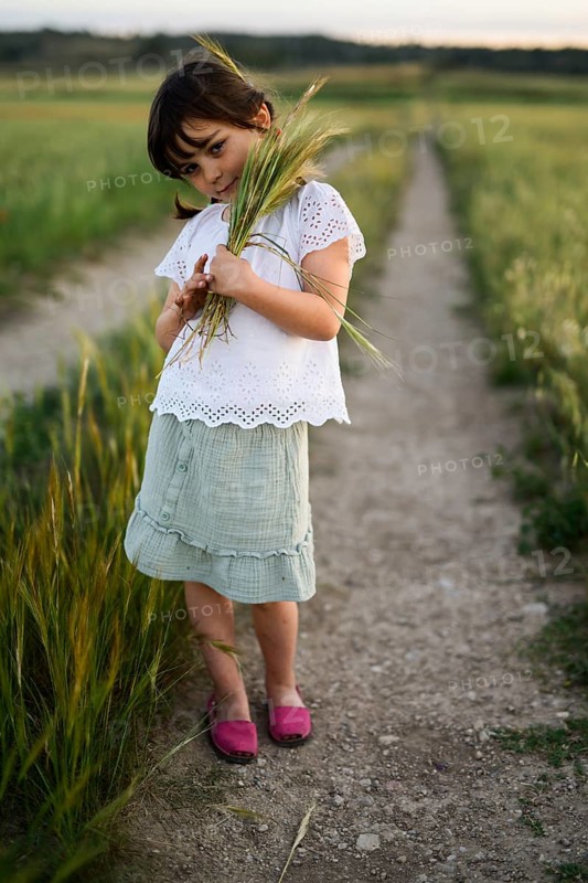 A young girl, dressed in a white blouse and green skirt, gently holds a bunch of wheat while standing on a rural path. Her contemplative expression and the soft evening light create a peaceful scene in nature
