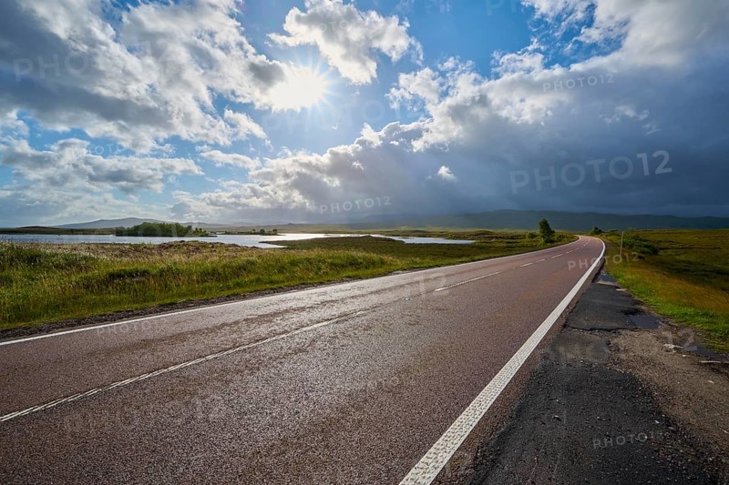 Long road with beautiful sunlight, flanked by greenery and expanses of water, cloudy on the horizon, Rannoch Moor, Bridge of Orchy, Scotland, United Kingdom, Great Britain