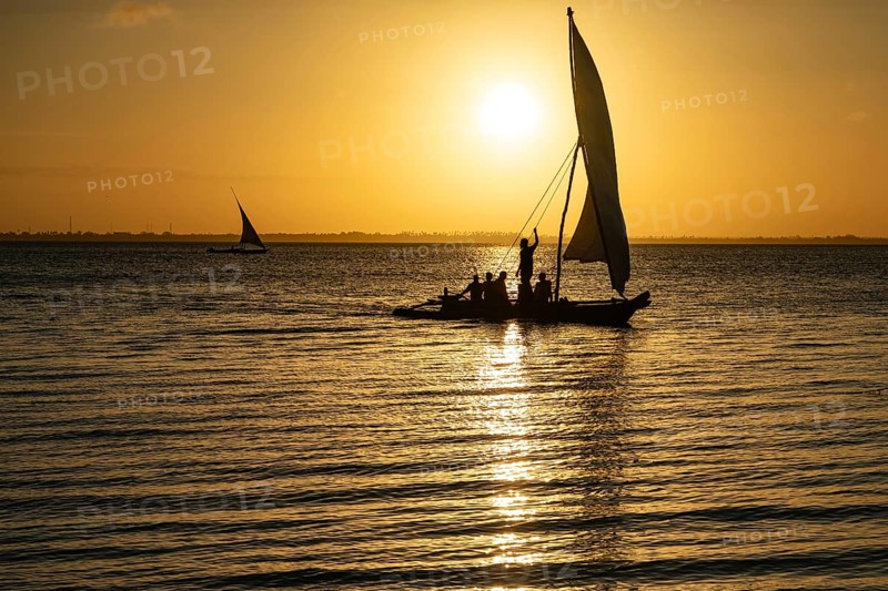 Silhouette of a traditional dhow sailing boat with people on board against a golden sunset over the ocean in Zanzibar