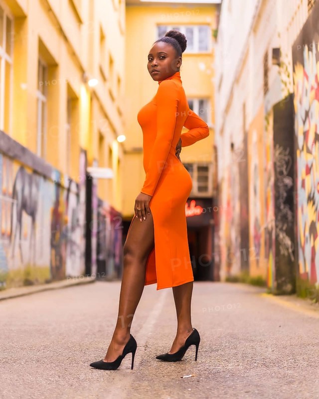 Fashionable urban style with a black African girl in an orange dress and black heels on a city street. Standing pose with seductive gaze