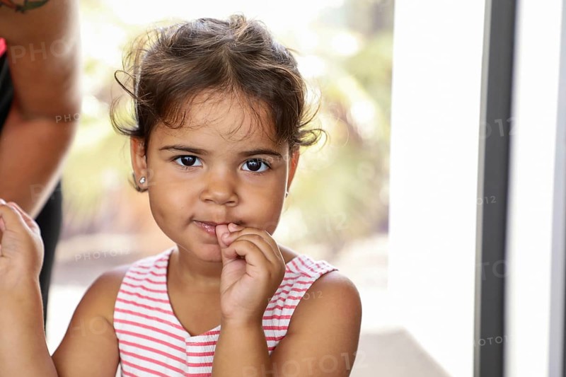 A young child in a striped dress stares curiously at the camera, surrounded by soft natural light. Her thoughtful expression captures a moment of innocence and wonder