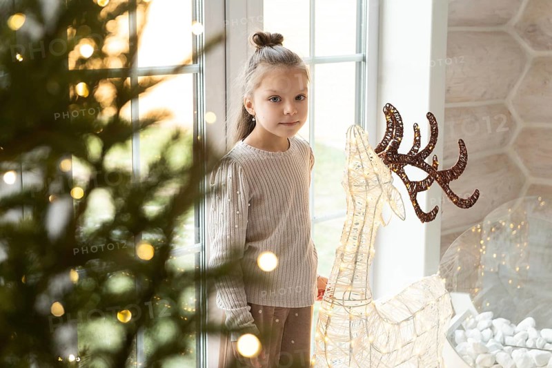 A young girl stands by a window adorned with Christmas lights, looking at camera, next to a decorative light-up reindeer