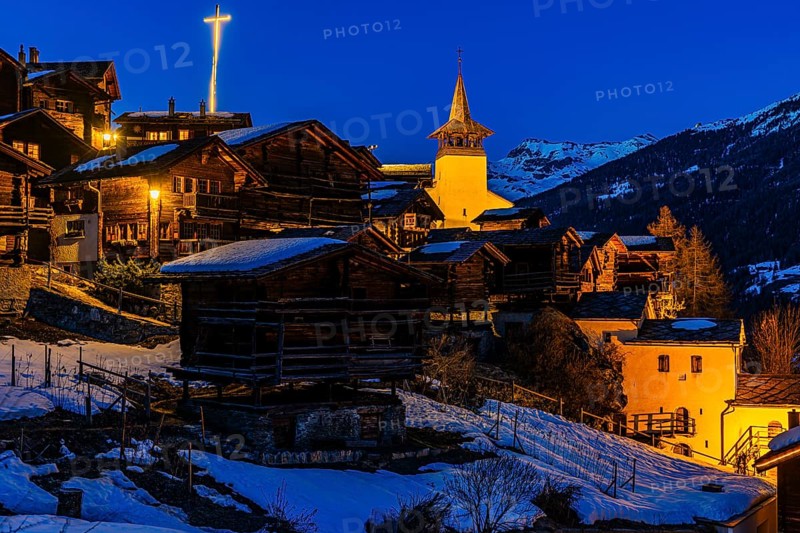 Snow-covered roofs of old farmhouses and barn storage, in the back the illuminated cross and the illuminated church tower of the Saint Theodule church, night view, historic town center, Grimentz, Val d'Anniviers, Valais Alps, Canton of Valais, Switzerland