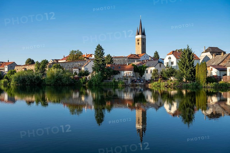 A wide panoramic view of the historic town of Telc with its colorful buildings and church towers reflected in the calm surface of a pond under a clear blue sky in the Czech Republic