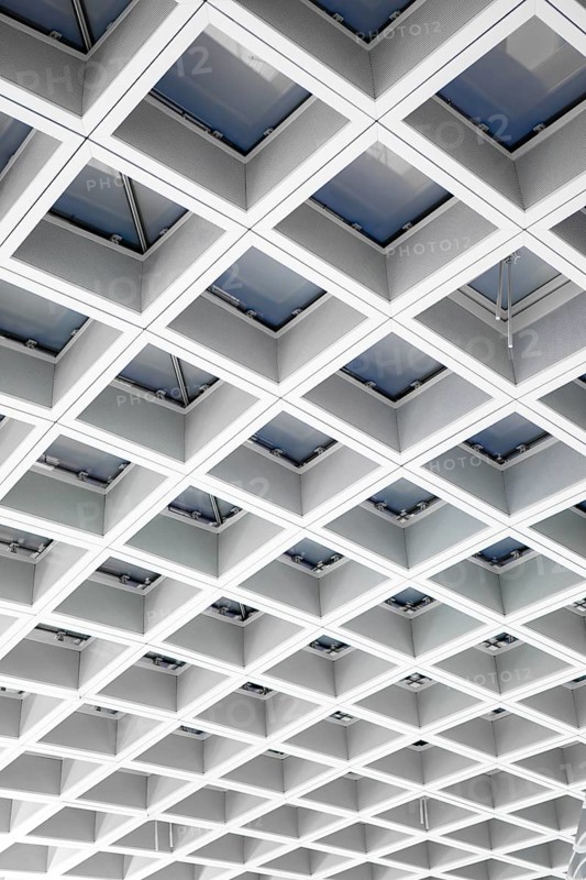 A modern airport ceiling features a geometric grid of skylights, creating a pattern of light and shadow. This architectural design enhances the travel experience with style