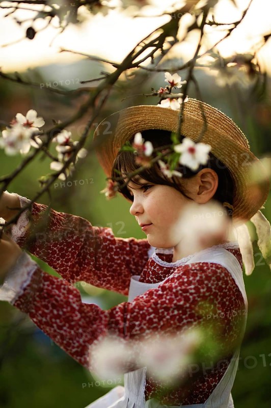 A young girl in a red dress and straw hat admires the blossoms of an almond tree. The scene captures the essence of childhood, nature, and the tranquility of spring