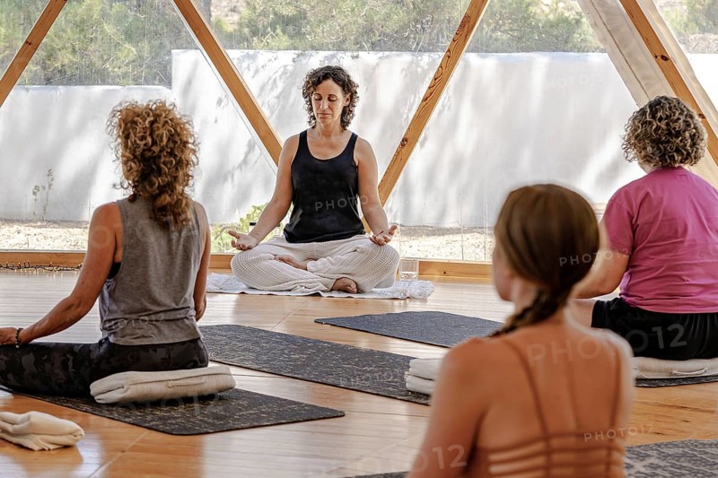 Woman guides a peaceful meditation session at a studio. With her eyes closed, she sits cross-legged, while clients sit facing away, creating a tranquil atmosphere