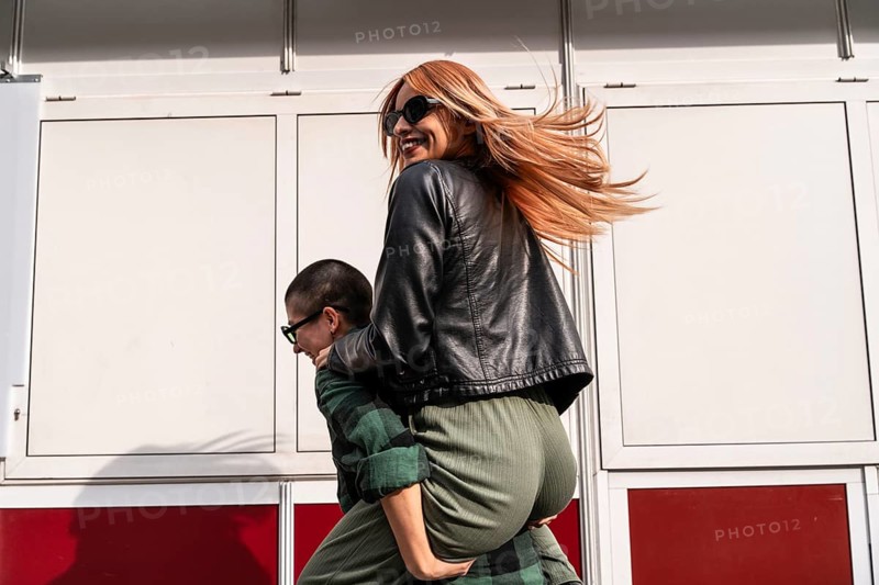 A joyful Colombian lesbian couple shares a fun piggyback ride, laughing and enjoying the moment. Their stylish outfits, sunglasses, and carefree energy reflect love, happiness, and adventure