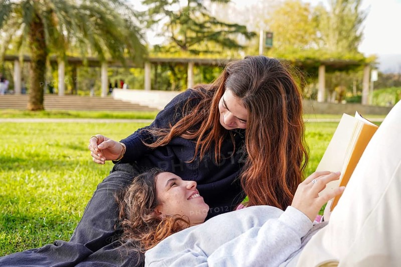 A romantic young women couple, part of the LGBTQ+ community, lovingly shares a moment while reading a book in a lush park. They are lying on the grass, one looking at the book while the other gazes adoringly at her partner