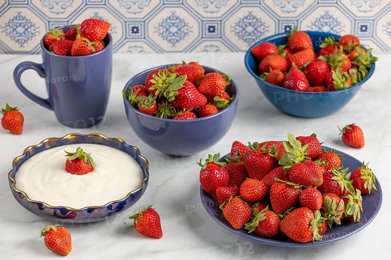 Fresh strawberries arranged in blue crockery next to a bowl of yoghurt on a marble plate