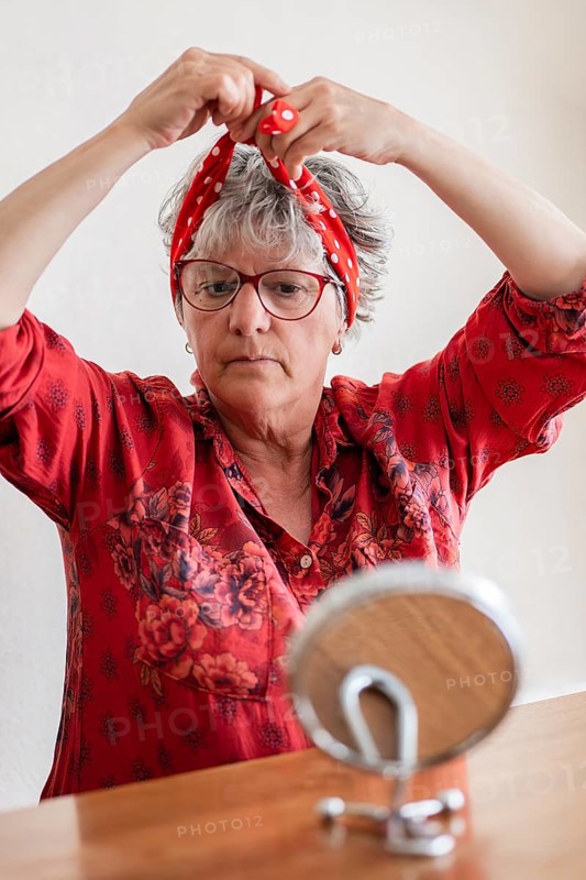 A senior woman with gray hair and glasses ties a red polka dot headscarf in front of a mirror. Dressed in a red floral blouse, she focuses on creating a stylish accessory