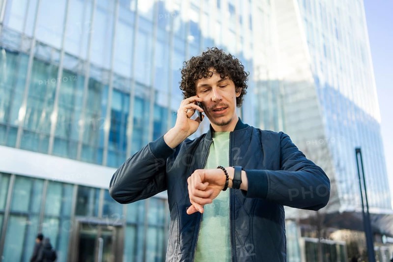 A businessman stands in front of a glass building, talking on his smartphone while checking the time on his smartwatch, symbolizing productivity and multitasking
