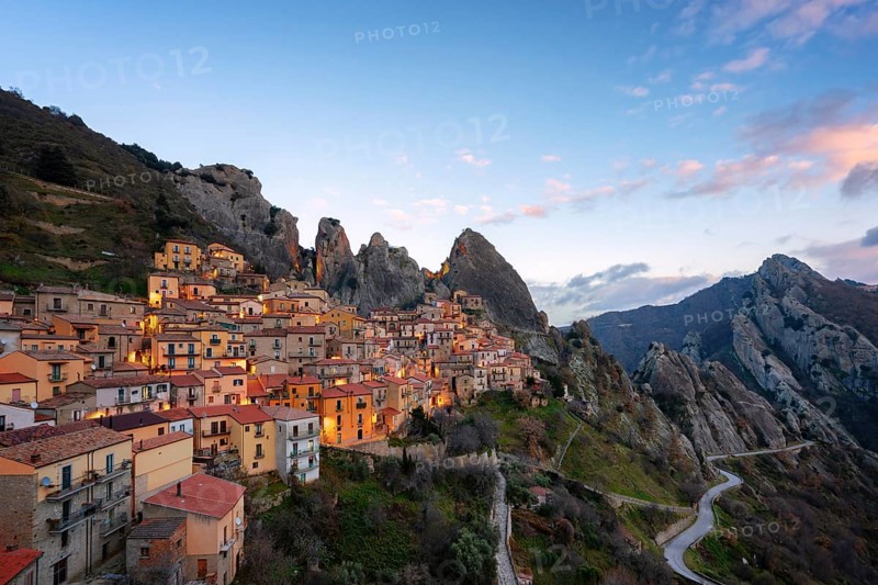 The enchanting village of Castelmezzano, nestled in the Lucanian Dolomites, glows under a vibrant sunset. Its stone houses blend seamlessly with the rugged mountain landscape