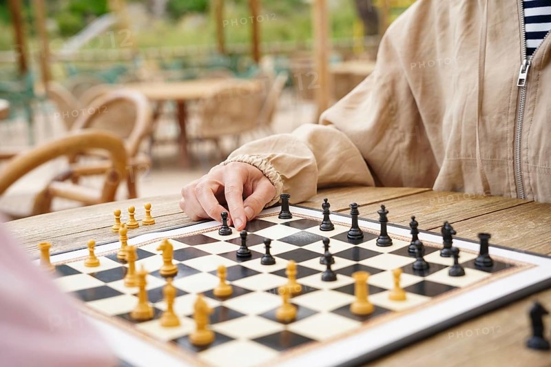 A close-up view of a chess game being played outdoors on a wooden table The chessboard is set with black and white pieces, surrounded by a relaxed, sunny atmosphere