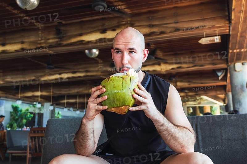 A bald man sips from a green coconut while relaxing indoors on his vacation in Bali. This casual, holiday-themed scene captures the essence of leisure and exotic refreshment