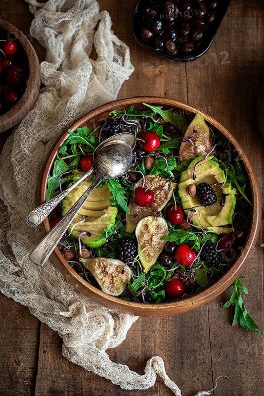 A vibrant salad featuring fresh figs, avocado, cherries, and blackberries on a bed of arugula, sprinkled with almonds in a wooden bowl, set on a rustic wooden table