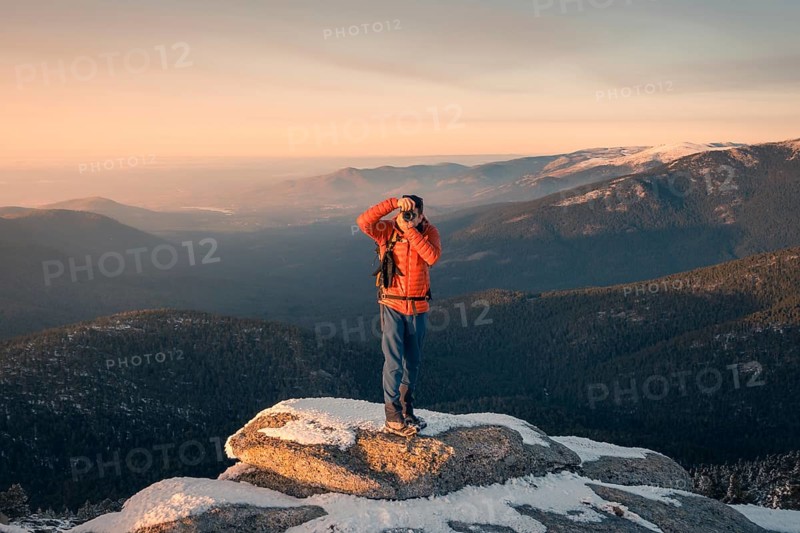 A photographer stands atop a snow-dusted peak at Siete Picos, Guadarrama Mountains, capturing the vast landscape of Madrid during sunset. Breathtaking winter scene