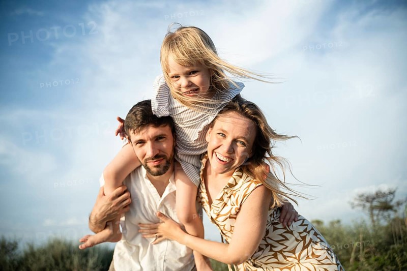 A joyful family enjoys a sunny day at the beach in Italy. The child sits on the father's shoulders as the mother smiles beside them, capturing a moment of happiness