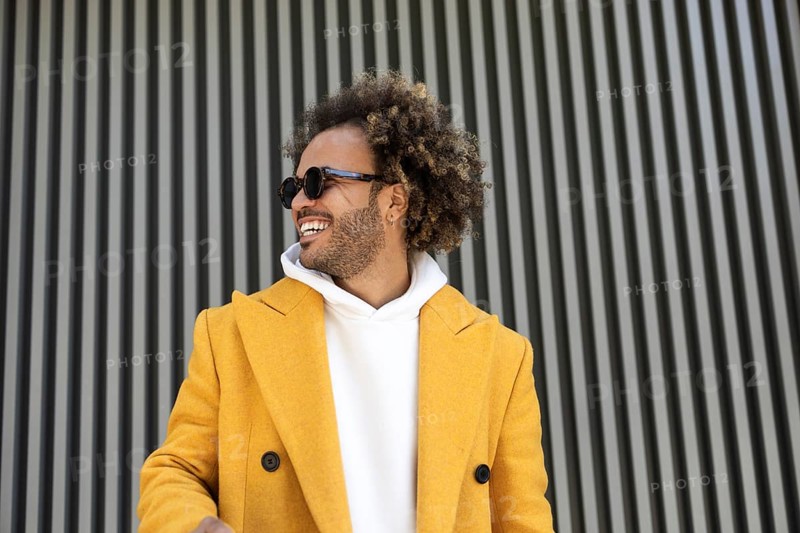 An afro man wearing a vibrant yellow coat and sunglasses, standing confidently against a modern, striped backdrop, exuding style and a carefree attitude