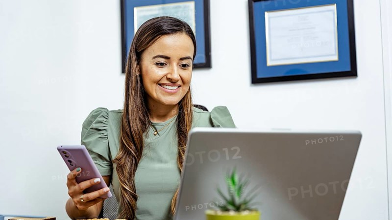 A nutritionist in a modern office setting is engaged in an online consultation using a laptop and smartphone. Diplomas are displayed in the background, creating a professional atmosphere