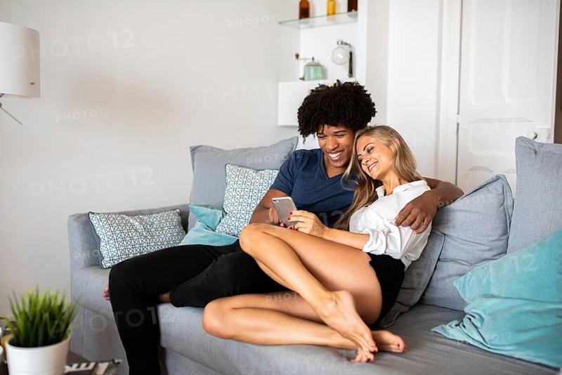 A couple relaxes on a comfortable sofa, smiling as they look at a smartphone. The living room setting is cozy, filled with soft cushions and neutral tones