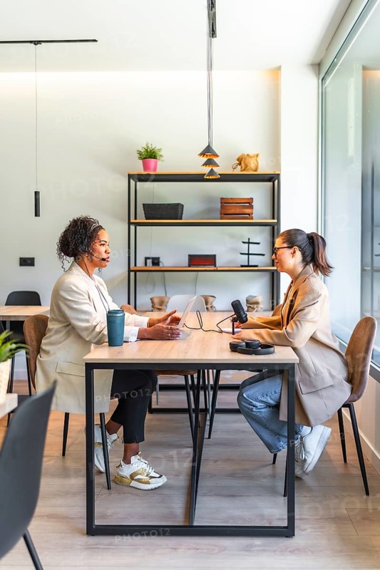 Two businesswomen are recording a podcast in a modern office, using a microphone and laptop