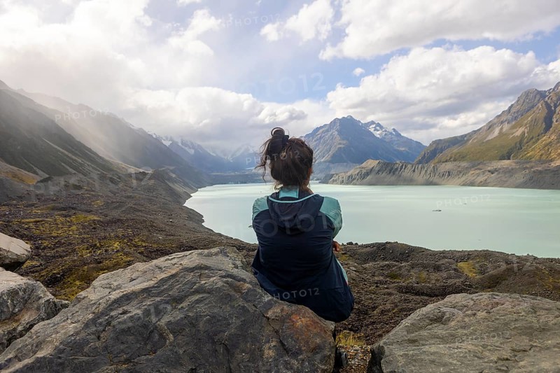A woman sits on a rocky outcrop, taking in the breathtaking autumn scenery of Mount Cook, New Zealand. The highlands are bathed in soft, seasonal hues beneath a cloudy sky