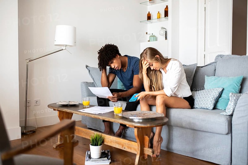 A couple sits on a couch, looking worried as they go over financial documents with a calculator. Nearby, glasses of orange juice sit on the wooden table in a cozy room