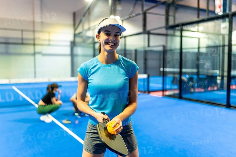 Young smiling woman holding a pickleball paddle and ball, standing on a vibrant blue court in an indoor sports facility, enjoying an active recreation