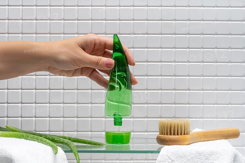 Cropped unrecognizable woman dispensing aloe vera gel from a bottle over a bathroom sink, with body care accessories nearby