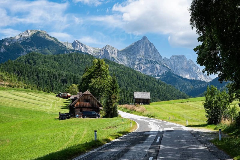 A winding road leads past traditional wooden houses and green meadows toward the sharp, towering peaks of the Austrian Alps under a blue sky in Austria
