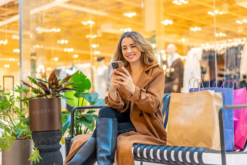 Woman in a trench coat sitting on a bench, looking at her phone and smiling, with shopping bags next to her in a modern mall environment, enjoying online shopping and retail therapy