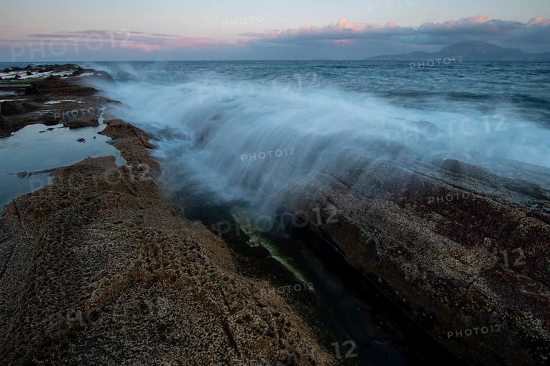 Waves crash against rocky shores under a vivid sunset sky in Tarifa, Cadiz. The vibrant colors and dynamic motion create a dramatic seascape